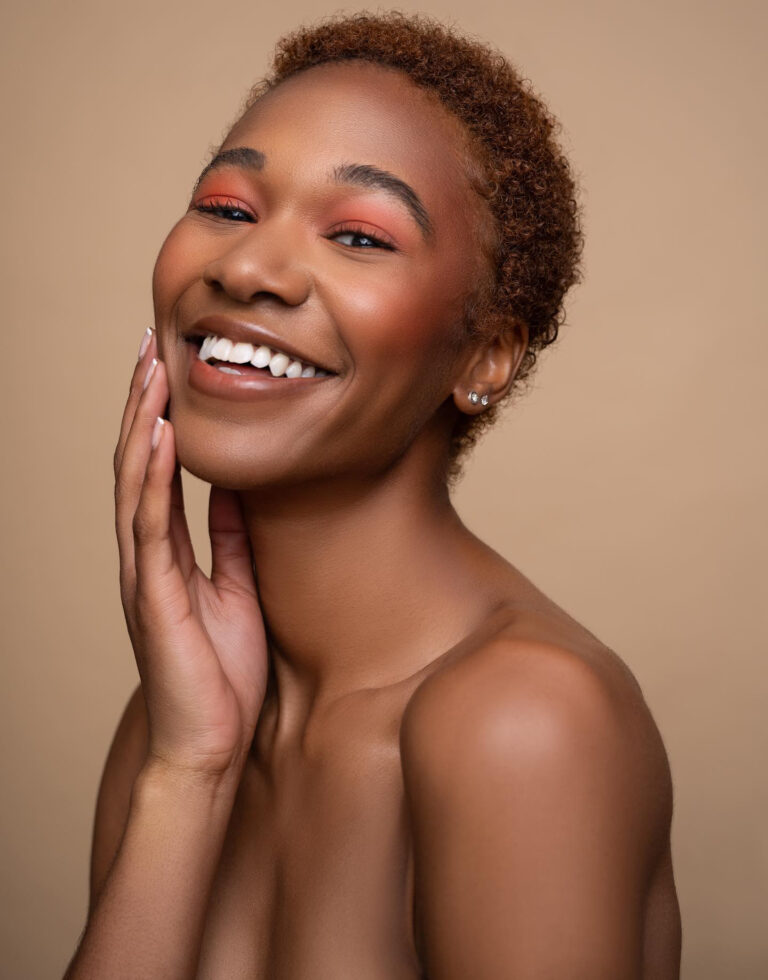 Beauty headshot of black woman smiling and holding chin in soft orange colored makeup.