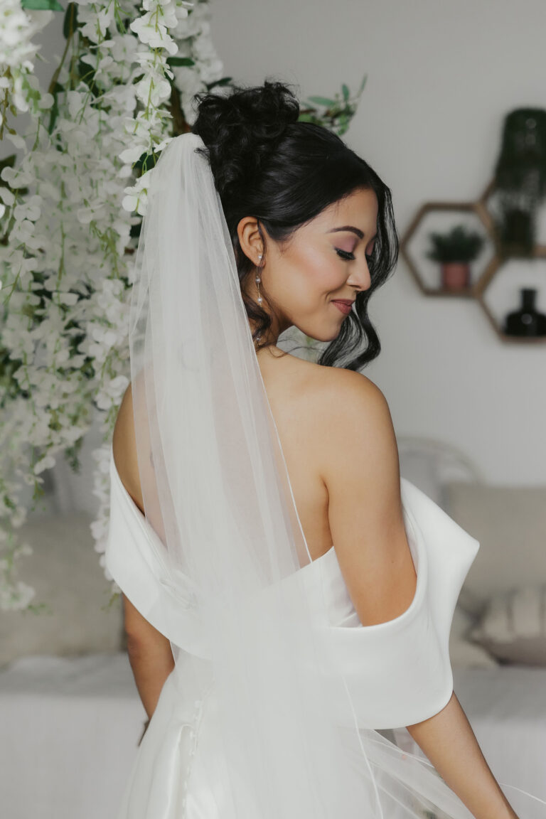 Bride in white gown and veil smiling with back facing camera and showing bridal makeup.