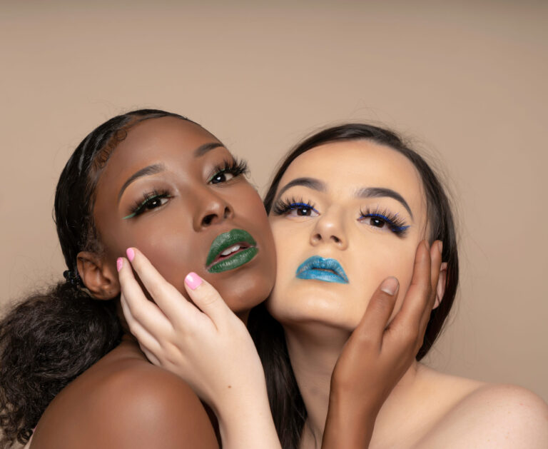 Black model and green lipstick and white model with blue lipstick cheek to cheek, holding one another's opposite cheek.