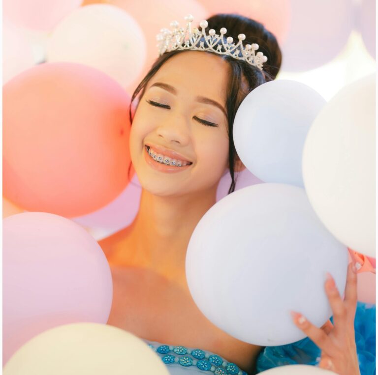 Young girl dressed as a princess, smiling joyfully while surrounded by pastel-colored balloons, wearing a tiara and light blue dress.